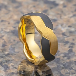 May include: A gold and black wedding band with a unique, geometric design. The band is made of two different metals, with a black metal band interwoven with a gold band.