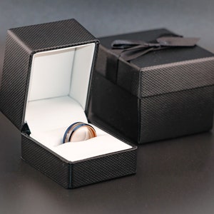 May include: A black ring box with a white interior, open to reveal a silver and blue ring with a black band.