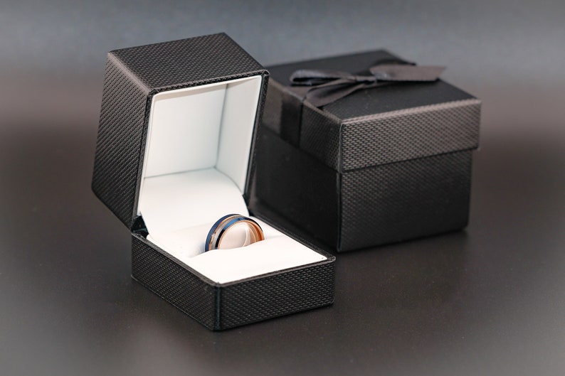 May include: A black ring box with a white interior, open to reveal a silver and blue ring with a black band.