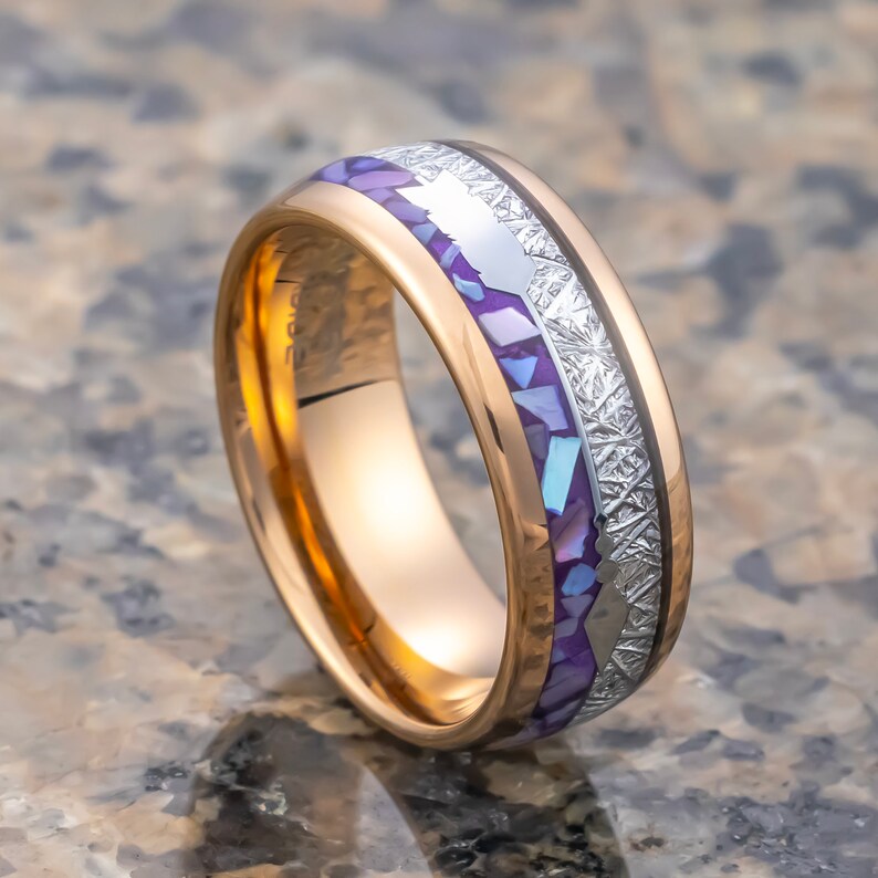 Puede incluir: Un anillo de carburo de tungsteno de oro rosa con una incrustaci&oacute;n de color p&uacute;rpura y blanco. La incrustaci&oacute;n presenta un patr&oacute;n de peque&ntilde;as formas irregulares y una banda plateada.