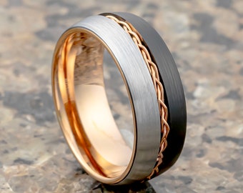 May include: A black and silver tungsten carbide ring with a rose gold braided band.