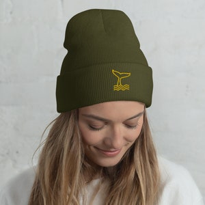 Op de afbeelding: Een donkergroene beanie met een geborduurd geel walvisstaartontwerp. De beanie is gemaakt van een zachte, gebreide stof.