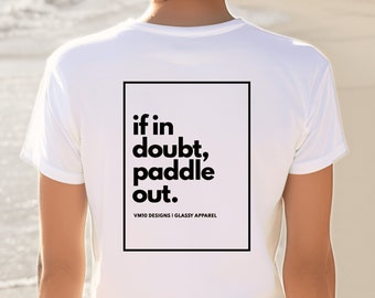Puede incluir: Una camiseta blanca con el texto "if in doubt, paddle out" en un marco rectangular negro. La camiseta también incluye el texto "VM10 DESIGNS | GLASSY APPAREL". La imagen está tomada en una playa.