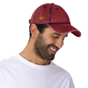 Puede incluir: Un hombre con una gorra de béisbol roja desgastada con un logotipo de aleta de tiburón bordado en amarillo que dice "WHO" en la parte delantera. Lleva una camiseta blanca.
