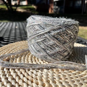 May include: A skein of yarn in a light grey colour with silver and black flecks. The yarn is sitting on a woven wicker surface.