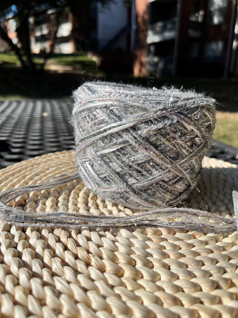 May include: A skein of yarn in a light grey colour with silver and black flecks. The yarn is sitting on a woven wicker surface.