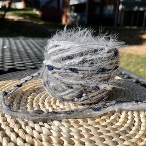 May include: A skein of yarn with a fuzzy texture in shades of grey, white, and blue. The yarn is wrapped around a woven wicker mat.