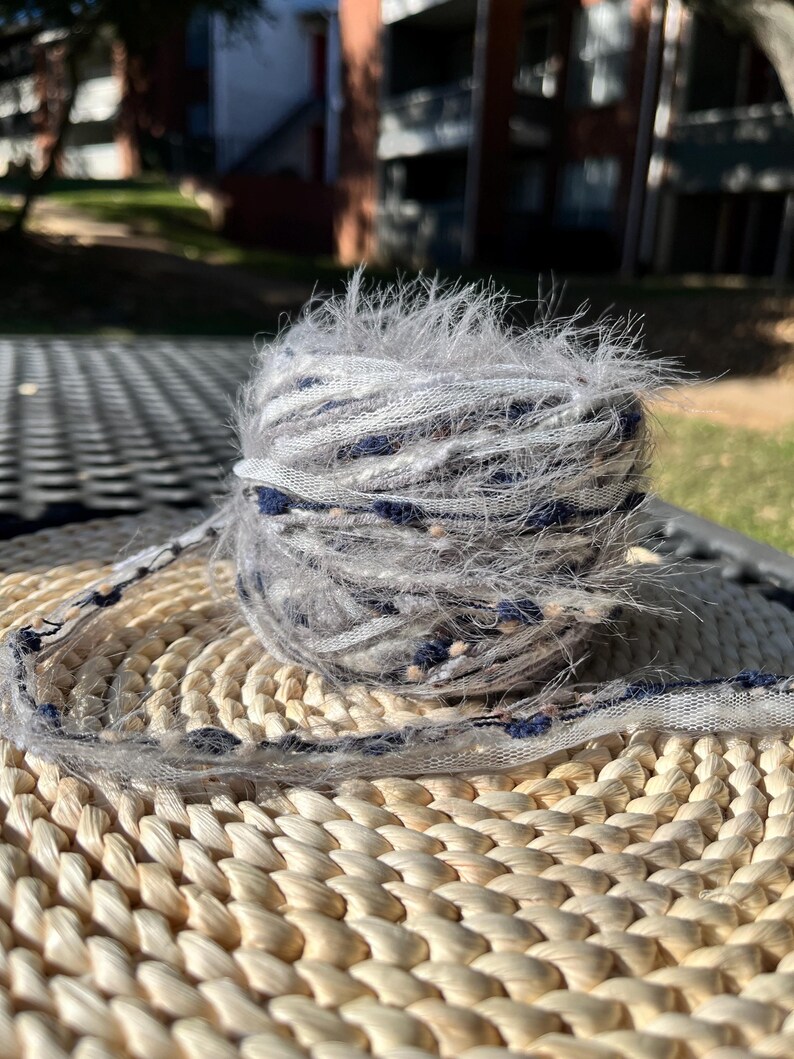 May include: A skein of yarn with a fuzzy texture in shades of grey, white, and blue. The yarn is wrapped around a woven wicker mat.