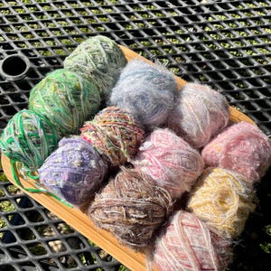 May include: A collection of colorful yarn balls in a variety of textures, including fuzzy, sparkly, and striped. The yarn is arranged in a wooden tray on a black metal surface.