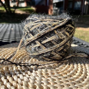 May include: A ball of black and gold yarn with a fuzzy texture. The yarn is sitting on a woven wicker surface.