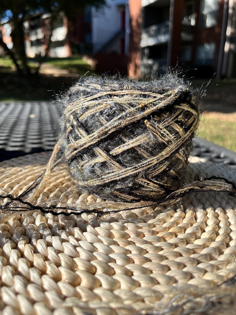 May include: A ball of black and gold yarn with a fuzzy texture. The yarn is sitting on a woven wicker surface.