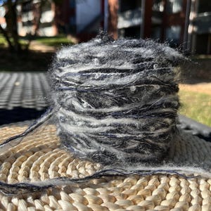 May include: A skein of black and white yarn with a fuzzy texture. The yarn is wound into a ball shape and is sitting on a woven straw mat.
