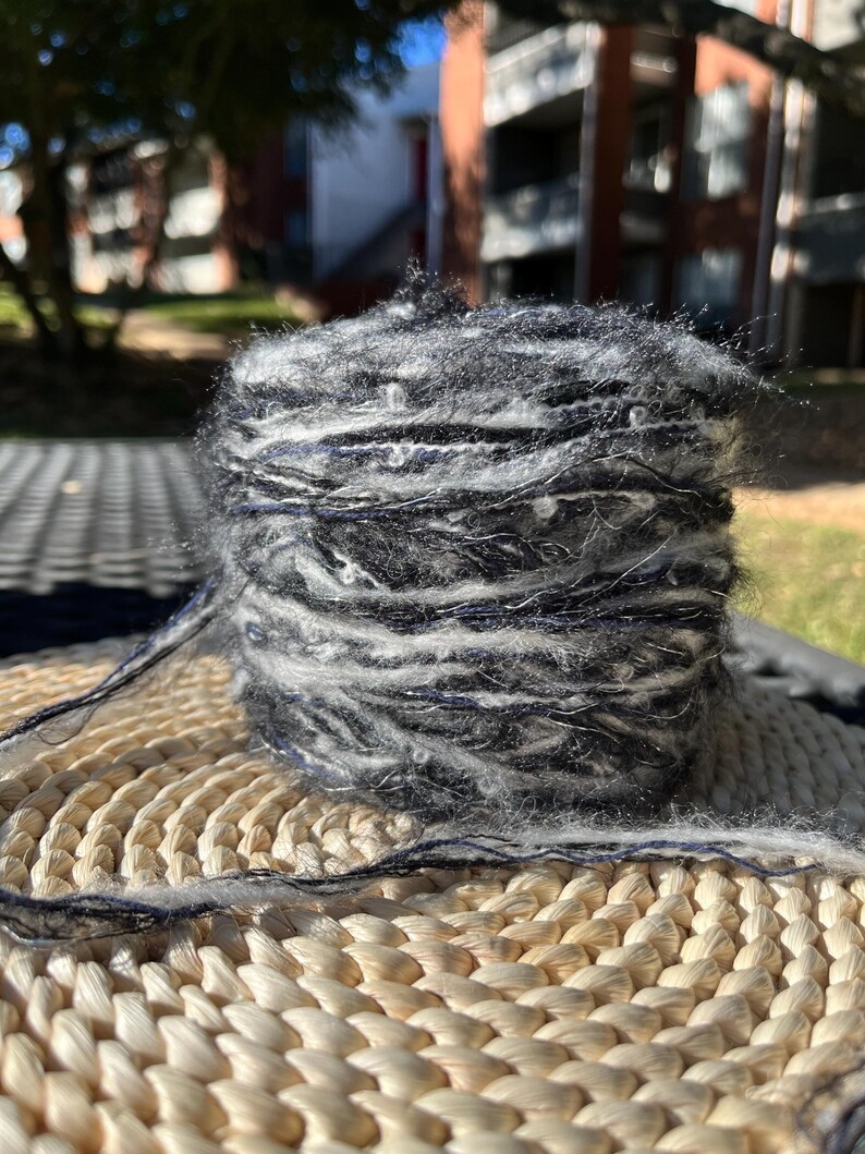 May include: A skein of black and white yarn with a fuzzy texture. The yarn is wound into a ball shape and is sitting on a woven straw mat.