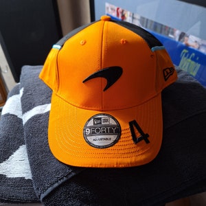 May include: Orange and black baseball cap with the McLaren logo. The cap features the New Era 9FORTY adjustable design and the number 4. The cap is made of a durable material and has a curved peak.