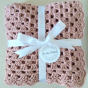 May include: A dusty rose crochet blanket, neatly folded and tied with a white satin ribbon. A circular tag with the text "On the Prairie" is attached. The blanket's open weave pattern is visible.