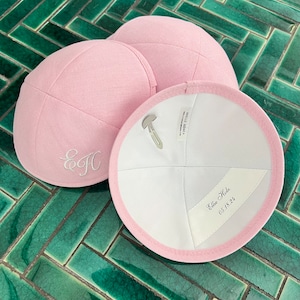 K&ouml;nnte beinhalten: Zwei rosa Kippahs, eine mit den Initialen "E.J." in wei&szlig;er Stickerei und die andere mit dem Text "Ellie Hale 05.18.22" in schwarzer Tinte.