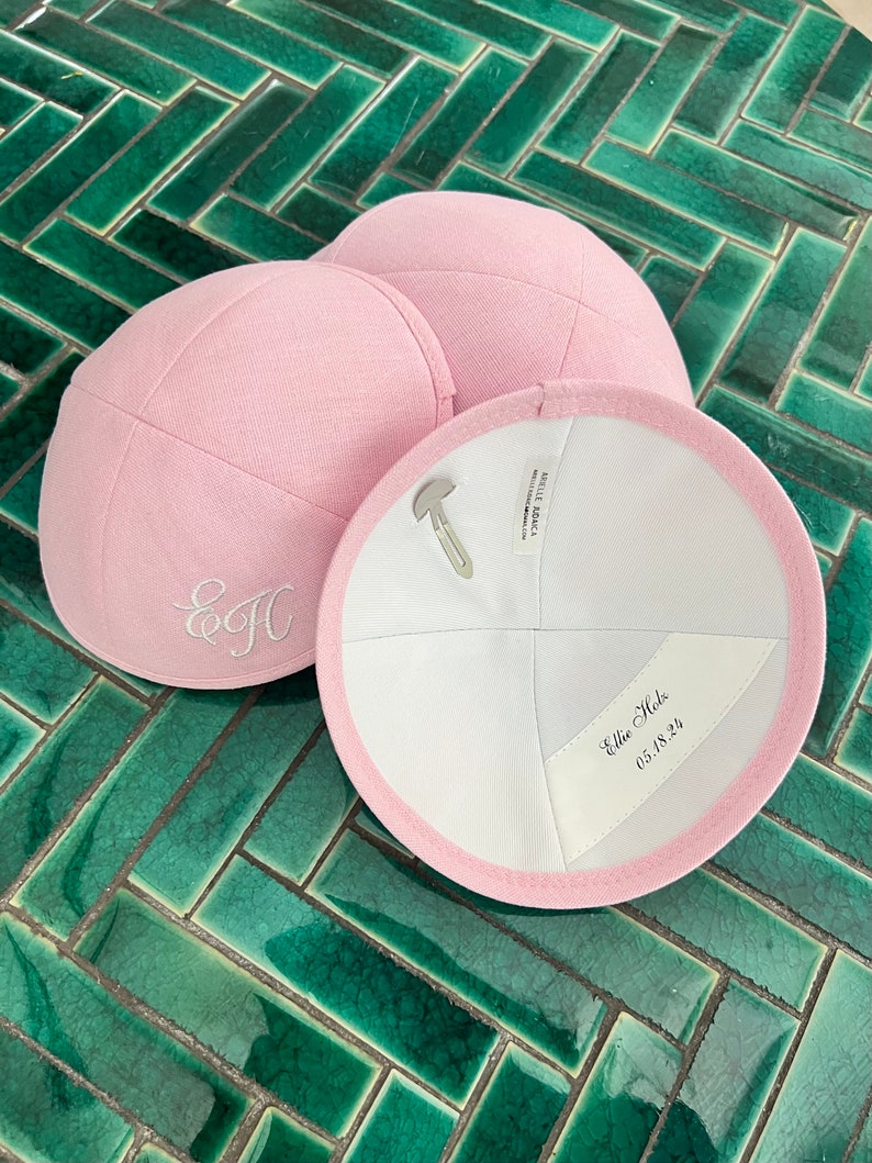 K&ouml;nnte beinhalten: Zwei rosa Kippahs, eine mit den Initialen "E.J." in wei&szlig;er Stickerei und die andere mit dem Text "Ellie Hale 05.18.22" in schwarzer Tinte.