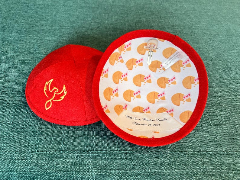 K&ouml;nnte beinhalten: Rote Filz-Kippa mit einem goldenen gestickten Ph&ouml;nix-Design. Die Innenseite der Kippa ist mit einem wei&szlig;en Stoff ausgekleidet, der ein Muster aus Gl&uuml;ckskeksen mit rosa Herzen zeigt. Die Auskleidung enth&auml;lt auch ein wei&szlig;es Etikett mit dem Text "With Love, Penelope Landis, September 28, 2023."