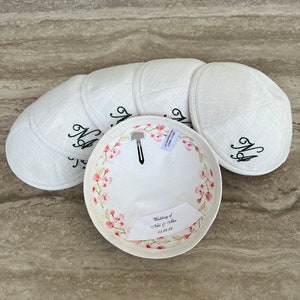 K&ouml;nnte beinhalten: Sechs wei&szlig;e Kippahs mit Blumendesigns und dem Text "Wedding of Noa & Allie 05.28.23" auf der Innenseite.