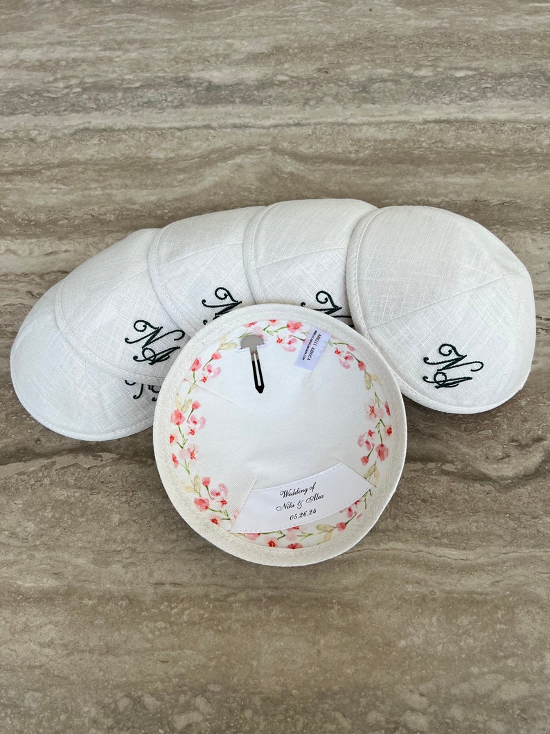 K&ouml;nnte beinhalten: Sechs wei&szlig;e Kippahs mit Blumendesigns und dem Text "Wedding of Noa & Allie 05.28.23" auf der Innenseite.