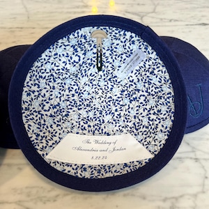 K&ouml;nnte beinhalten: Eine marineblaue Kippa mit einer wei&szlig;en Blumenfutter. Die Kippa hat ein wei&szlig;es Etikett mit dem Text "The Wedding of Alexandria and Jordan 8.22.24".