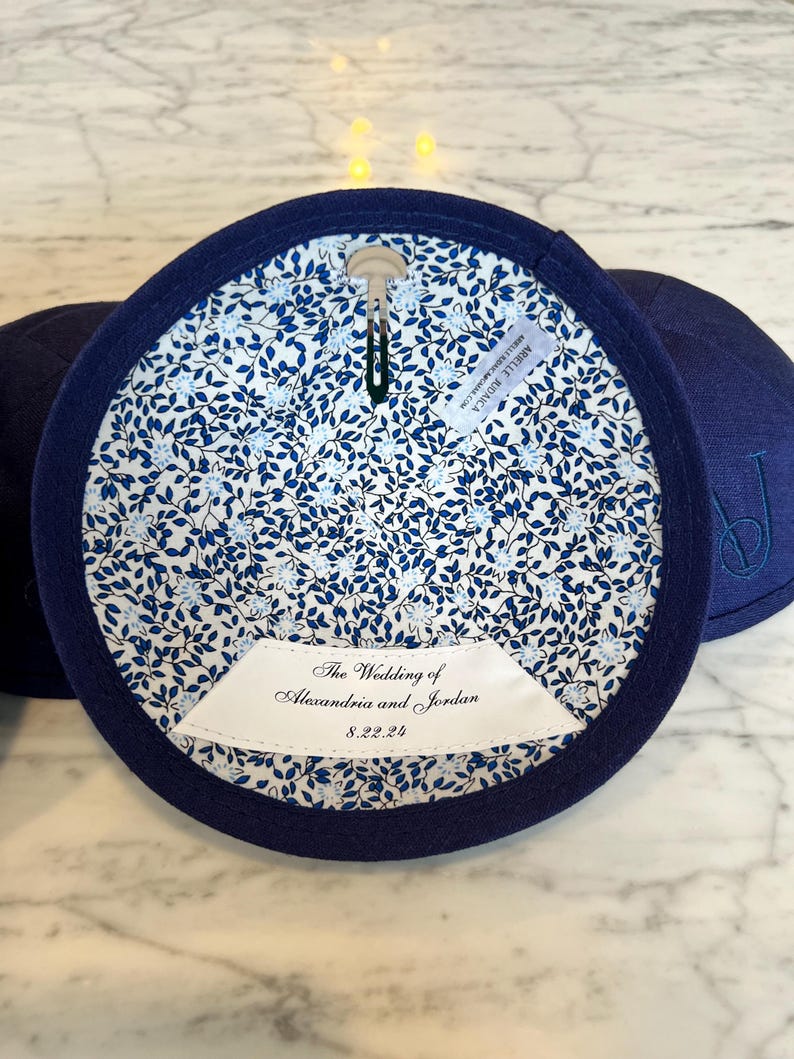 K&ouml;nnte beinhalten: Eine marineblaue Kippa mit einer wei&szlig;en Blumenfutter. Die Kippa hat ein wei&szlig;es Etikett mit dem Text "The Wedding of Alexandria and Jordan 8.22.24".