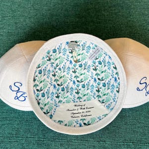 K&ouml;nnte beinhalten: Drei wei&szlig;e Kippahs mit blauer Stickerei. Die Kippa in der Mitte hat ein florales Futter und ein Etikett mit der Aufschrift "Hochzeit von Alexander & Beth Conner, 23. September 2023, Valencia, Kalifornien."