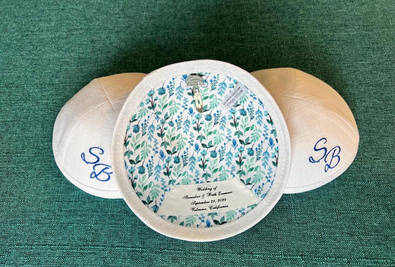 K&ouml;nnte beinhalten: Drei wei&szlig;e Kippahs mit blauer Stickerei. Die Kippa in der Mitte hat ein florales Futter und ein Etikett mit der Aufschrift "Hochzeit von Alexander & Beth Conner, 23. September 2023, Valencia, Kalifornien."