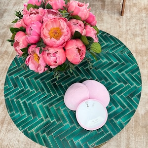 K&ouml;nnte beinhalten: Ein runder Tisch mit einer gr&uuml;nen gekachelten Oberfl&auml;che, auf dem ein Strau&szlig; rosa Pfingstrosen und drei rosa Kippahs stehen. Zwei der Kippahs stehen auf dem Tisch und eine liegt auf der anderen.