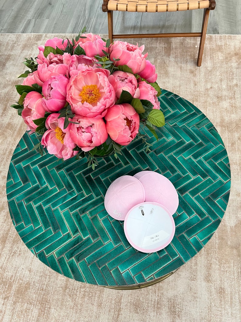 K&ouml;nnte beinhalten: Ein runder Tisch mit einer gr&uuml;nen gekachelten Oberfl&auml;che, auf dem ein Strau&szlig; rosa Pfingstrosen und drei rosa Kippahs stehen. Zwei der Kippahs stehen auf dem Tisch und eine liegt auf der anderen.
