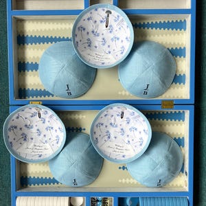 K&ouml;nnte beinhalten: Eine blaue Holzkiste mit sechs blauen Kippahs darin. Die Kippahs haben ein wei&szlig;es Blumenmuster auf der Innenseite und die Initialen "J.B." auf der Au&szlig;enseite gestickt. Die Kiste enth&auml;lt auch wei&szlig;e und blaue Spielsteine.
