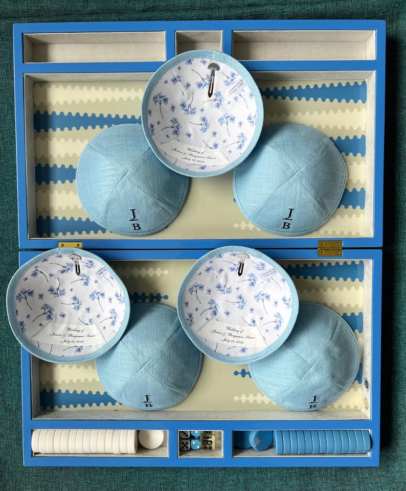 K&ouml;nnte beinhalten: Eine blaue Holzkiste mit sechs blauen Kippahs darin. Die Kippahs haben ein wei&szlig;es Blumenmuster auf der Innenseite und die Initialen "J.B." auf der Au&szlig;enseite gestickt. Die Kiste enth&auml;lt auch wei&szlig;e und blaue Spielsteine.
