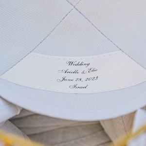 K&ouml;nnte beinhalten: Wei&szlig;er Stoff mit einem wei&szlig;en Etikett, auf dem "Wedding Arielle & Elie June 28, 2023 Israel" steht.