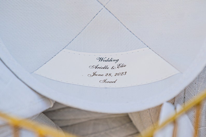 K&ouml;nnte beinhalten: Wei&szlig;er Stoff mit einem wei&szlig;en Etikett, auf dem "Wedding Arielle & Elie June 28, 2023 Israel" steht.