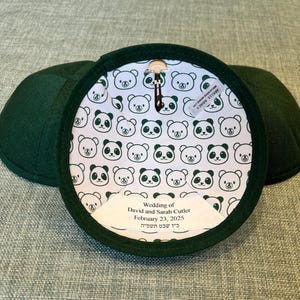 K&ouml;nnte beinhalten: Zwei gr&uuml;ne Kippahs mit einer wei&szlig;en Stoffauskleidung, die ein sich wiederholendes Muster aus schwarzen Pandab&auml;ren zeigt. Die Auskleidung enth&auml;lt auch ein wei&szlig;es Etikett mit dem Text "Wedding of David and Sarah Cutler February 23, 2025 כ'ו שבט תשפייה".