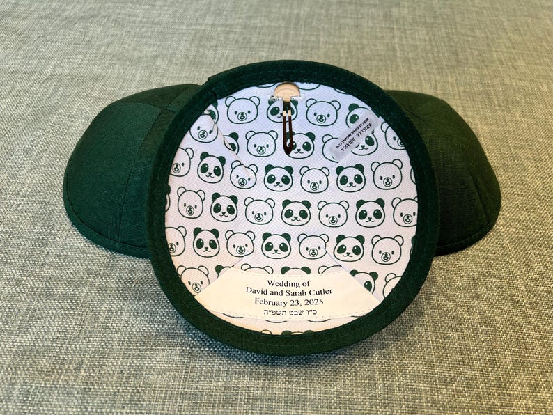 K&ouml;nnte beinhalten: Zwei gr&uuml;ne Kippahs mit einer wei&szlig;en Stoffauskleidung, die ein sich wiederholendes Muster aus schwarzen Pandab&auml;ren zeigt. Die Auskleidung enth&auml;lt auch ein wei&szlig;es Etikett mit dem Text "Wedding of David and Sarah Cutler February 23, 2025 כ'ו שבט תשפייה".