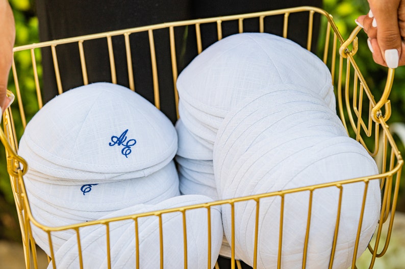 K&ouml;nnte beinhalten: Ein goldfarbener Drahtkorb gef&uuml;llt mit wei&szlig;en Kippahs aus Leinen, einige mit blauen Stickinitialen.