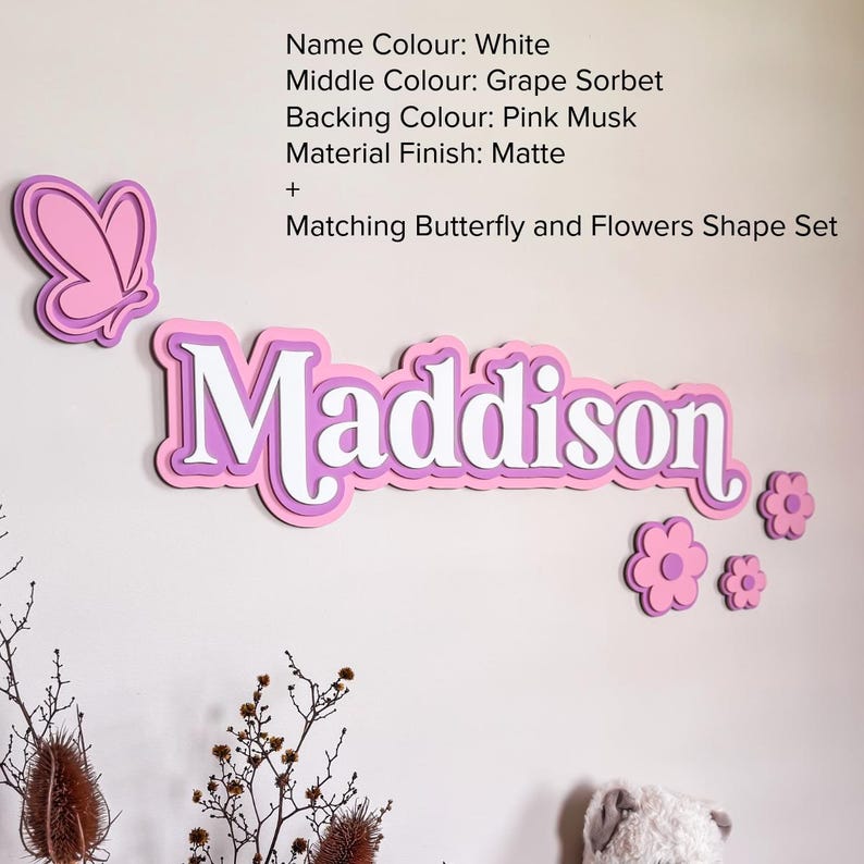 May include: A layered wall sign displaying the name Maddison in white, grape sorbet, and pink musk colors with a matte finish. The set includes a butterfly shape and three flower shapes. Text reads: Name Colour: White, Middle Colour: Grape Sorbet, Backing Colour: Pink Musk, Material Finish: Matte + Matching Butterfly and Flowers Shape Set.