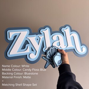 May include: A layered wall sign featuring the name Zylah in white, set against a candy floss blue middle layer and a bluestone backing. The sign includes a matching shell and starfish shape set with a matte finish and layered blue tones.