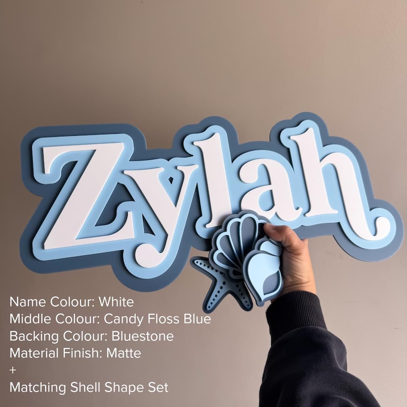 May include: A layered wall sign featuring the name Zylah in white, set against a candy floss blue middle layer and a bluestone backing. The sign includes a matching shell and starfish shape set with a matte finish and layered blue tones.