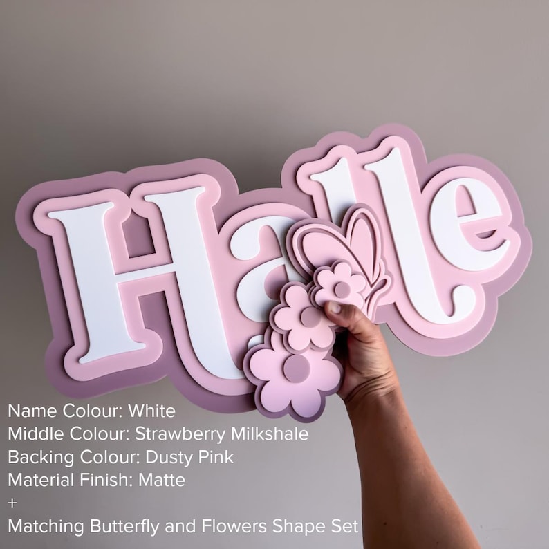 May include: A layered wall sign displaying the name Halle in white letters with a strawberry milkshake middle layer and a dusty pink backing. The sign features a matte finish and includes a matching butterfly and flower shape set attached.