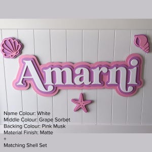 May include: A custom name sign featuring the name Amarni in white letters with a grape sorbet middle layer and a pink musk backing. The sign includes decorative shell and starfish accents in matching colors, all with a matte finish.