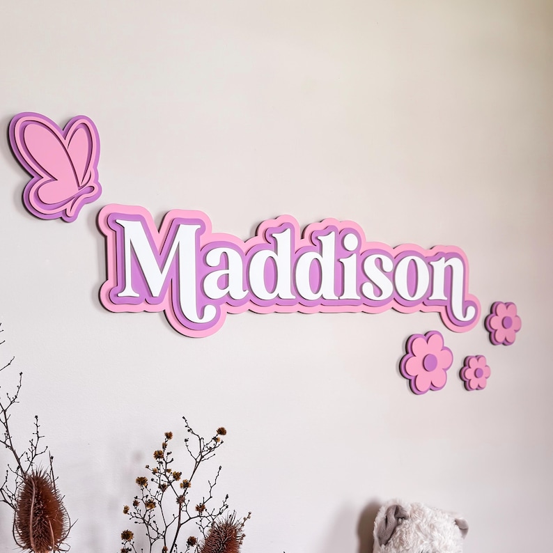 May include: A layered wall sign featuring the name Maddison in white letters with purple and pink outlines. The sign includes a pink butterfly icon to the left and three small pink flowers with purple centers positioned to the right of the name.