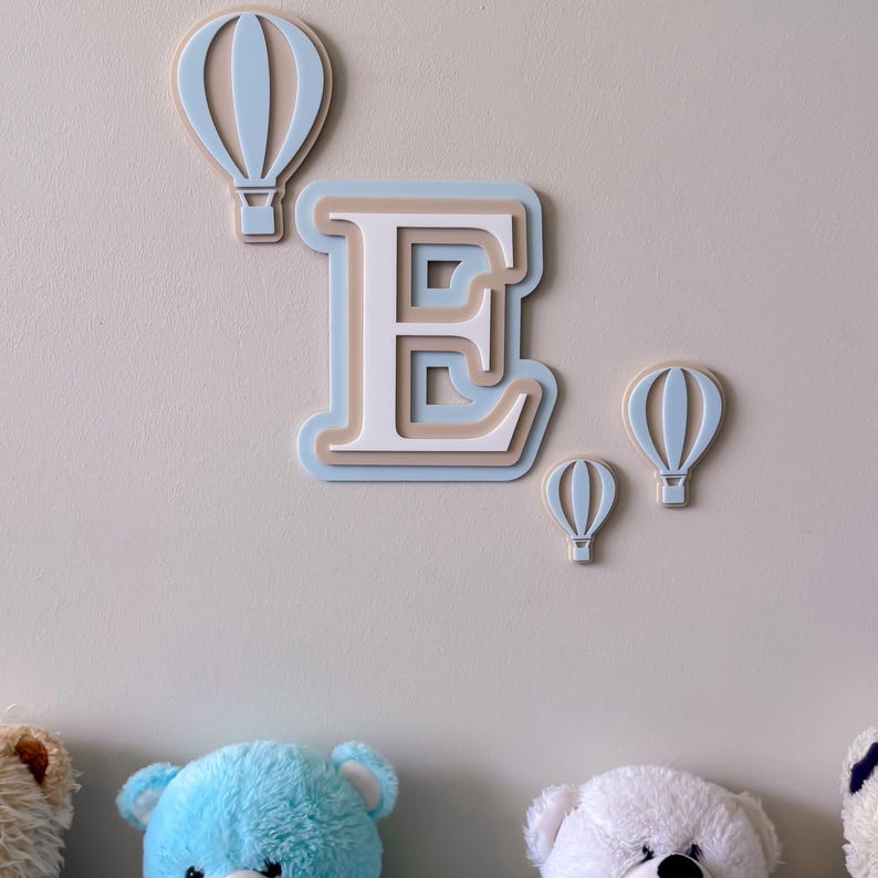 May include: A nursery wall decoration featuring a layered letter "E" in white, beige, and light blue, along with three hot air balloon cutouts in matching colors. The decor is mounted on a light gray wall.