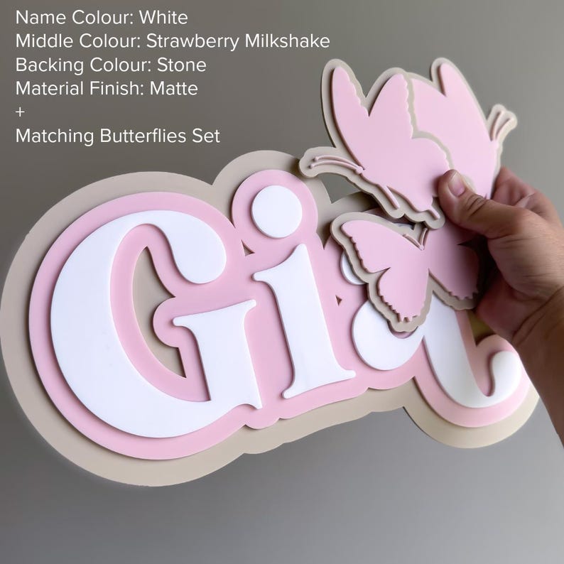 May include: A layered sign featuring the name Gia in white, set against a strawberry milkshake pink middle layer and a stone-colored backing. The matte finish sign includes a matching set of two pink butterfly cutouts attached to the right side.