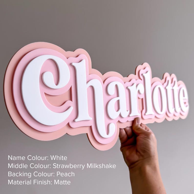 May include: A custom name sign featuring the name Charlotte in white, layered over a strawberry milkshake pink middle layer and a peach backing. The sign has a matte finish and displays the text: Name Colour: White, Middle Colour: Strawberry Milkshake, Backing Colour: Peach, Material Finish: Matte.