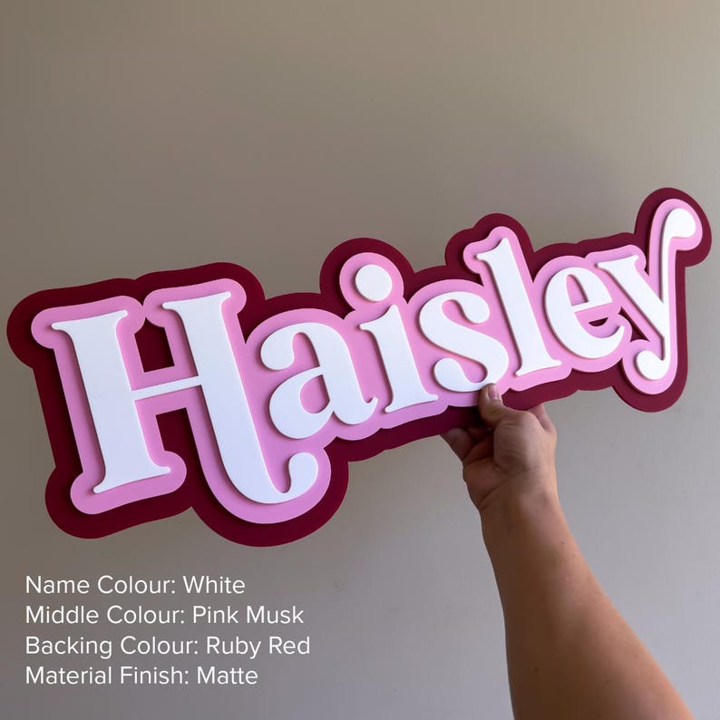May include: A custom name sign featuring the name Haisley in white lettering, layered over a pink musk middle border and a ruby red backing. The sign has a matte finish and displays the text: Name Colour: White, Middle Colour: Pink Musk, Backing Colour: Ruby Red, Material Finish: Matte.