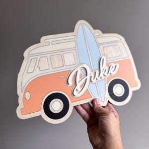 May include: Wooden wall art featuring a retro-style orange and cream van with a light blue surfboard. The name "Duke" is written in a 3D script. The van has black wheels and a roof rack.