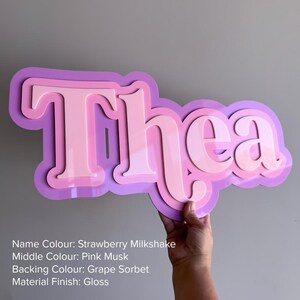 May include: A custom name sign featuring the name Thea in a layered, glossy design. The top layer is strawberry milkshake pink, the middle layer is pink musk, and the backing layer is grape sorbet purple. The sign has a smooth, reflective finish.