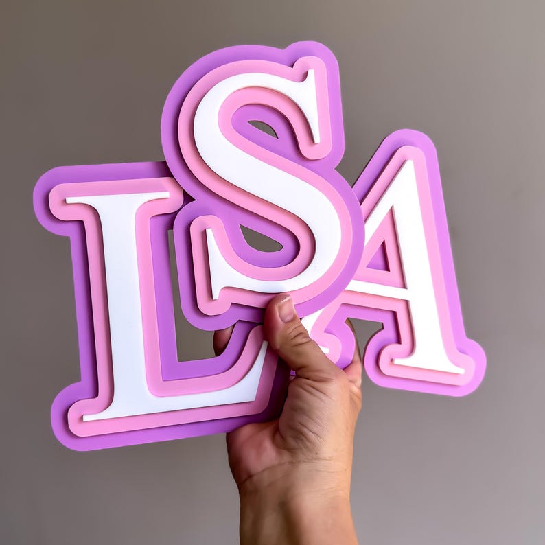 May include: Layered wooden letters spelling "LSA" in shades of purple, pink, and white. The letters are held against a neutral background. The letters are designed with a 3D effect, creating depth and dimension.